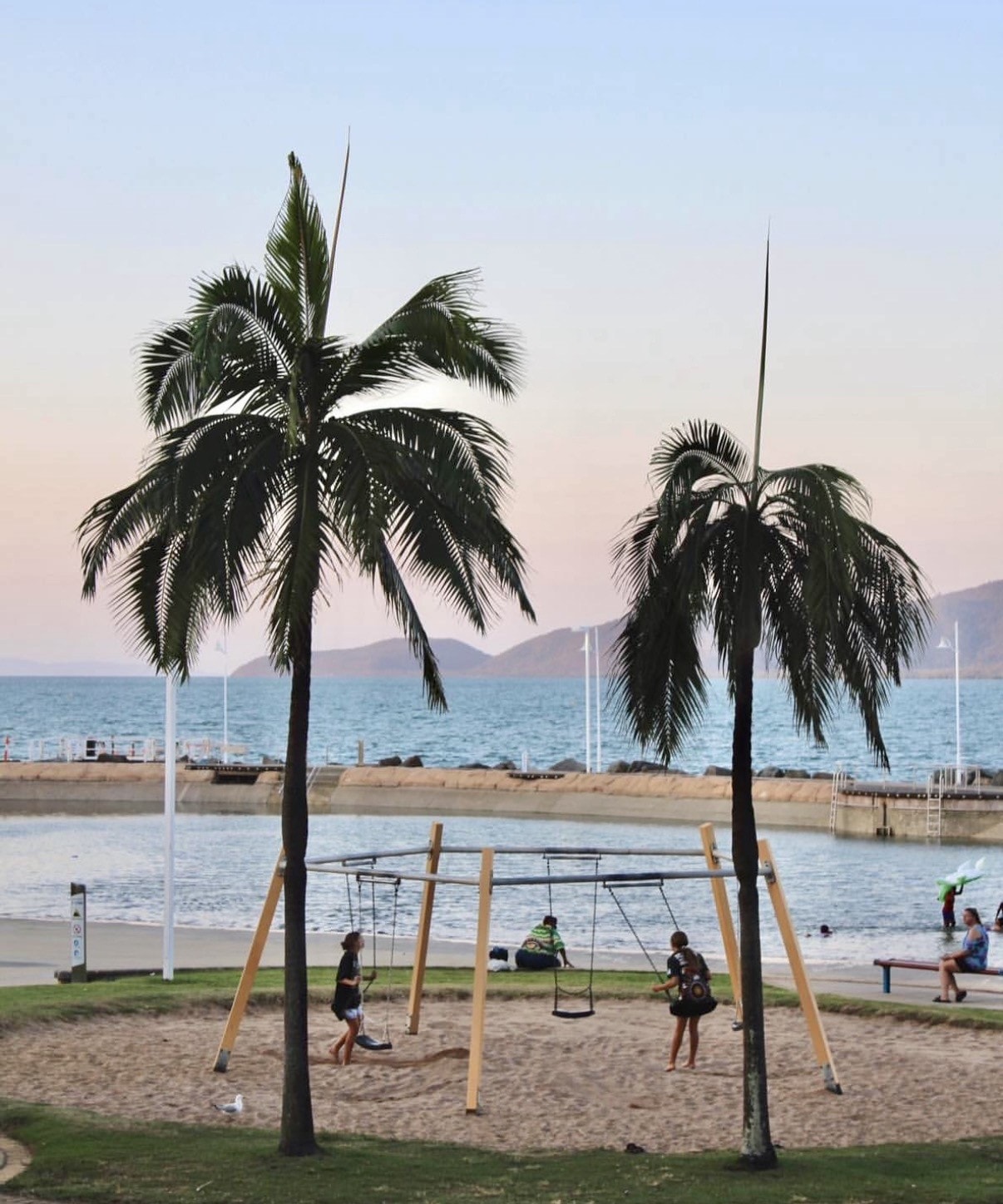 Children playing on swings at The Strand foreshore with palm trees, rockpool and mountain views at dusk